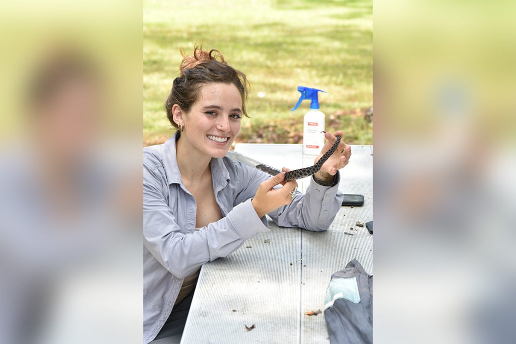 A person smiling and holding a small snake while sitting at a picnic table outdoors, with a spray bottle in the background.