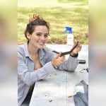 A person smiling and holding a small snake while sitting at a picnic table outdoors, with a spray bottle in the background.