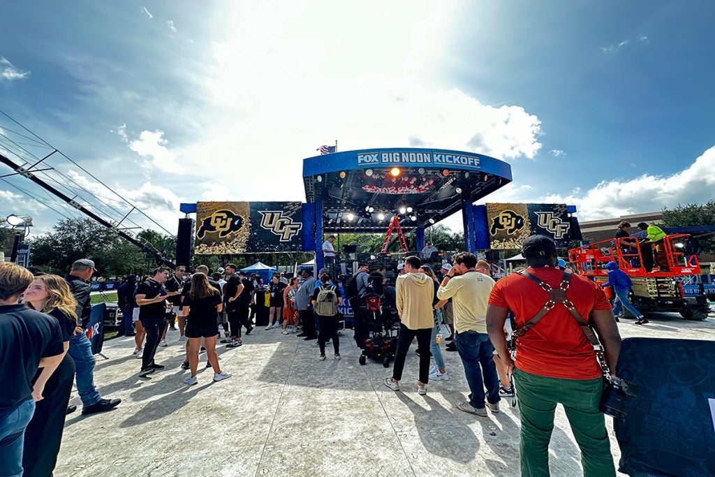 A crowd gathers near a stage with "Fox Big Noon Kickoff" banners. Logos of Colorado and UCF are displayed, surrounded by bright outdoor lighting under a partly cloudy sky.