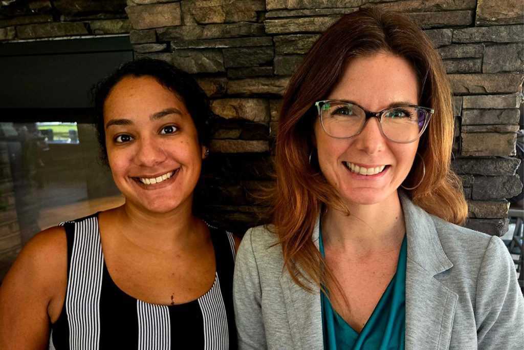 Two women smiling, standing side by side in front of a stone wall. One is wearing a striped top, and the other a blazer with glasses.