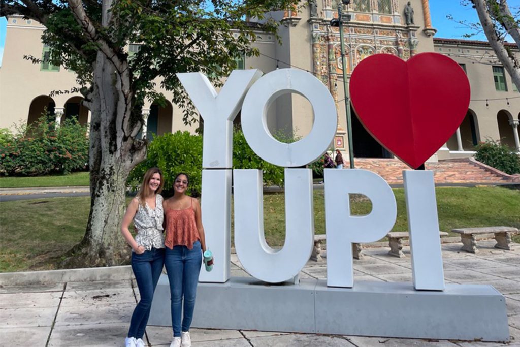 Two people smiling in front of a large "YO ❤️ UPI" sign with a decorative building and trees in the background.