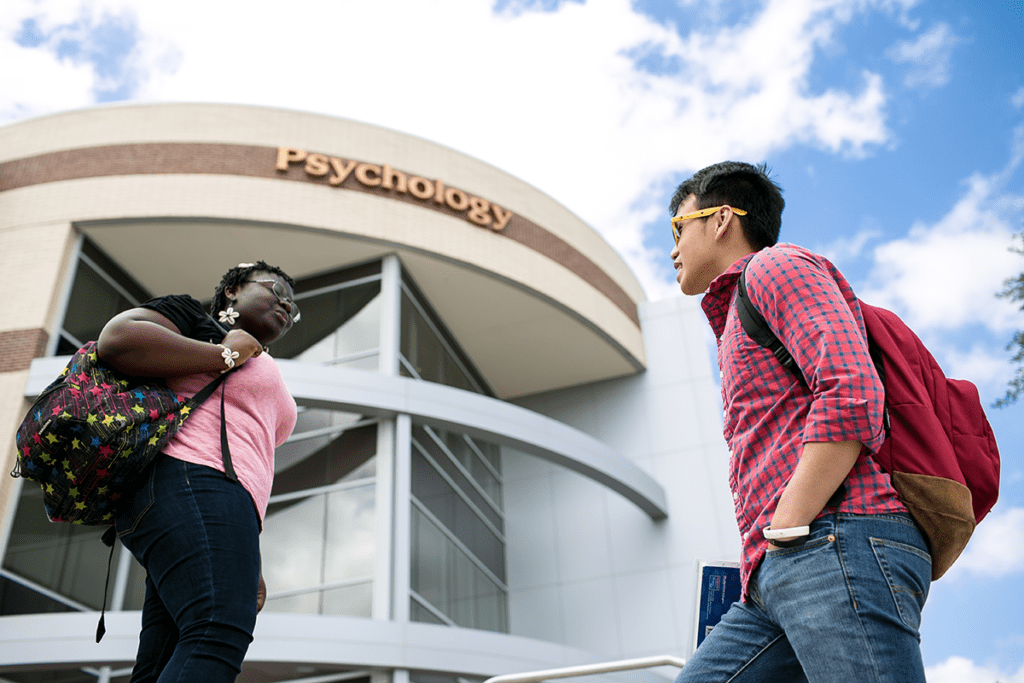 Two students with backpacks stand and talk in front of a building labeled "Psychology" under a blue sky.