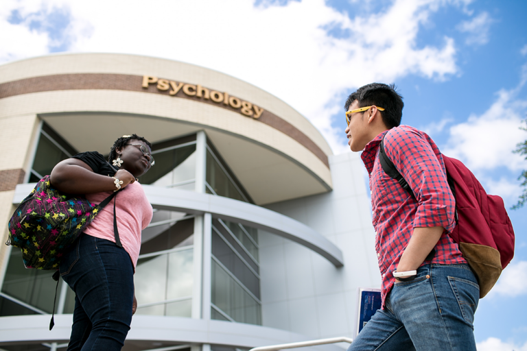 Two students with backpacks stand and talk in front of a building labeled 