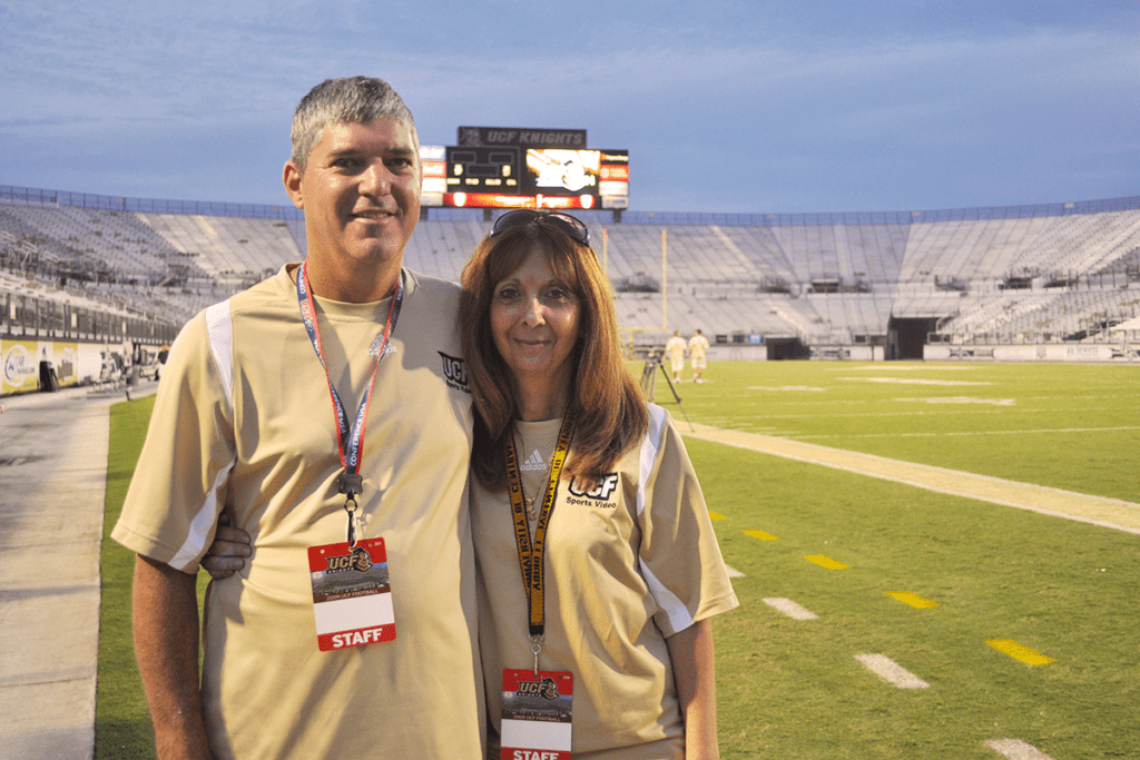 Two people in UCF staff shirts stand side by side on a football field with an empty stadium in the background.