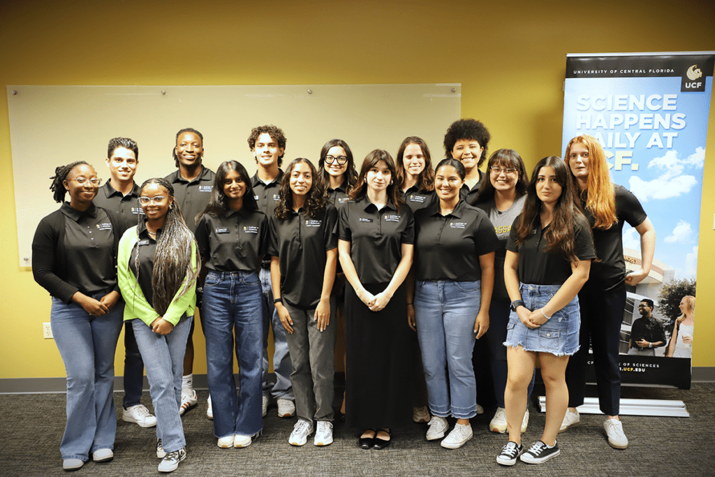 A diverse group of people wearing black polo shirts stand together in a room. A banner on the right reads, "Science happens daily at UCF.