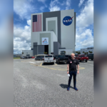 Person standing in a parking lot outside a large building with NASA and American flag logos.