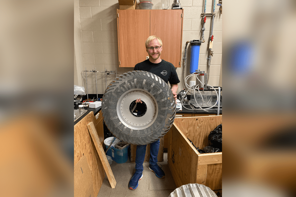 A person standing in a workshop holds a large, rugged tire in front of them, partially obscuring their body. Various equipment and tools are visible in the background.