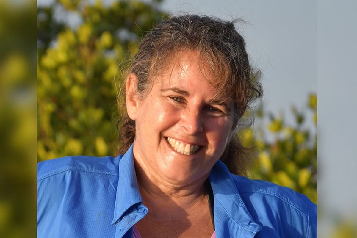 A person smiling outdoors, wearing a blue jacket. Trees are visible in the background.