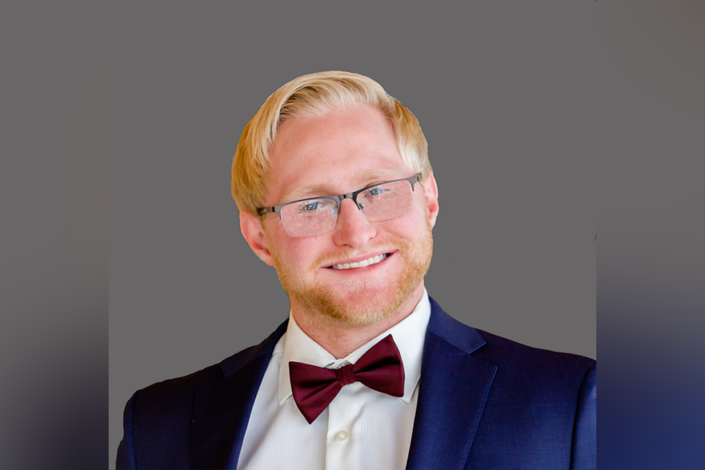 Man with glasses, blond hair, and a beard, wearing a dark suit, white shirt, and maroon bow tie, smiling against a gray background.