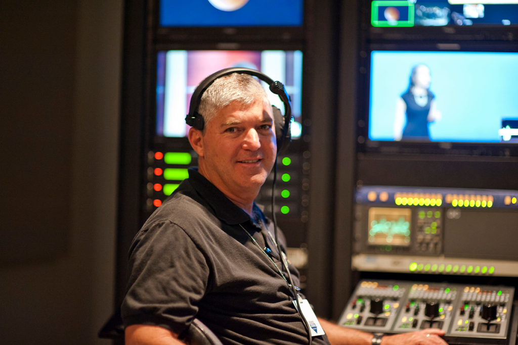 Man wearing a headset sits at a control panel with screens and equipment in a studio setting.