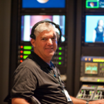 Man wearing a headset sits at a control panel with screens and equipment in a studio setting.