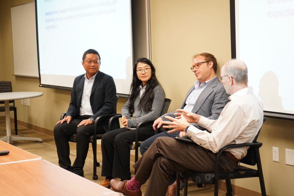 Four people sitting in chairs, engaged in a panel discussion in a conference room. A presentation is projected on the wall behind them.