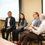 Four people sit in chairs discussing in a conference room with a presentation screen in the background.