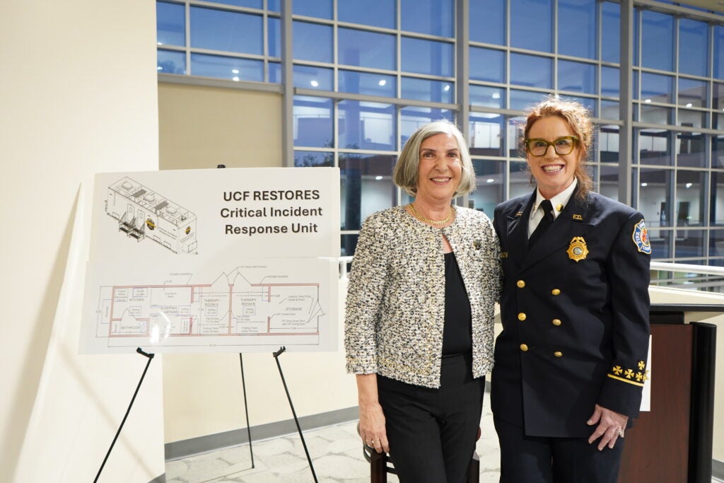 Two women stand indoors near a sign titled "UCF RESTORES Critical Incident Response Unit," featuring a diagram. One is in casual attire, the other in a uniform with a badge.