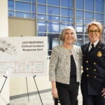 Two women stand indoors near a sign titled 