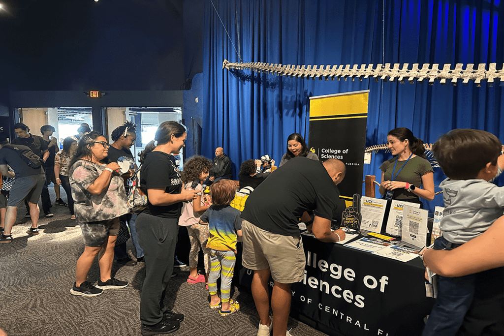 People gather around an informational booth for the College of Sciences at an indoor event. A banner and brochures are displayed on the table.