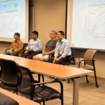 Four individuals seated in a conference room before a presentation screen displaying scientific graphs and text. Unoccupied chairs and tables in the foreground.