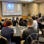 People seated at round tables attend a presentation in a conference room; a speaker stands at a podium while a screen displays the event.