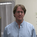 A man with glasses and light brown hair wearing a light blue button-down shirt stands indoors near a plant and a window with curtains.