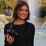 A young woman with short brown hair holds a camera and smiles while standing in front of a fountain in a park.