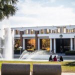 A modern building labeled "Millican Hall" stands behind a large fountain, with a few people sitting on the grass in the foreground.
