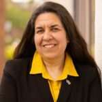 A woman with long dark hair, wearing a black blazer over a yellow shirt, stands smiling and leaning against a white wall.