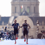 Two athletes in USA uniforms run side by side, connected by a tether, on a blue track with a blurred historic building in the background.