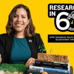A woman smiles while holding seagrass samples; a stack of books, plant models, and a sign reading “Research in 60s: How seagrass helps coastal ecosystems thrive” are visible.