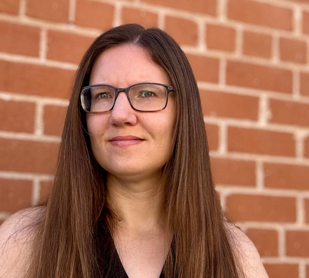 A woman with long brown hair and glasses stands in front of a brick wall, looking slightly to the side.