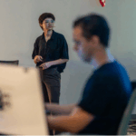 A person stands and looks to the side while another person sits blurred in the foreground, both in a modern indoor setting.