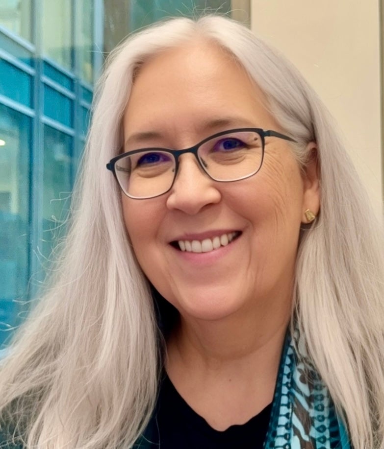 A woman with long gray hair and glasses is smiling, wearing a dark top, gold earrings, and a patterned scarf, standing indoors near a window with blue frames.