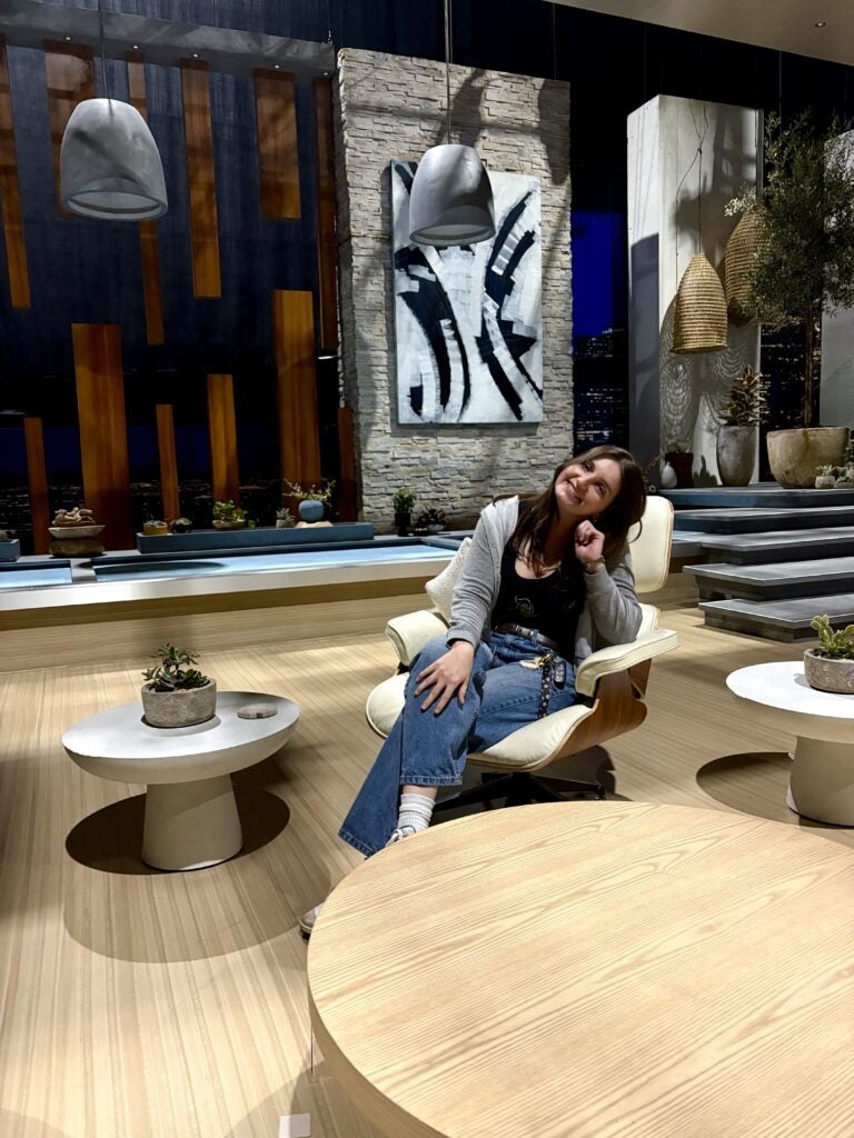 A woman sits and smiles on a modern chair in a stylish, contemporary indoor setting with plants, artwork, and decorative lighting.
