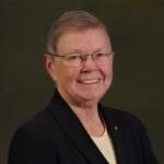 An older person with short gray hair and glasses, wearing a dark blazer and light shirt, smiles in front of a dark background.
