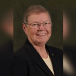 An older person with short hair, glasses, and a dark blazer over a light shirt, smiling in front of a dark background.