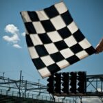 A black and white checkered flag is being waved above a racetrack with empty grandstands and signal lights visible in the background.