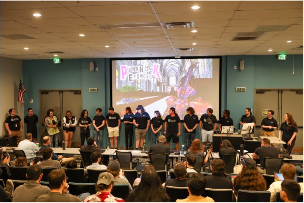 A group of people stands on stage facing an audience in a lecture hall; a screen behind them displays colorful "PUNK ROCK EXORCIST" graphics.