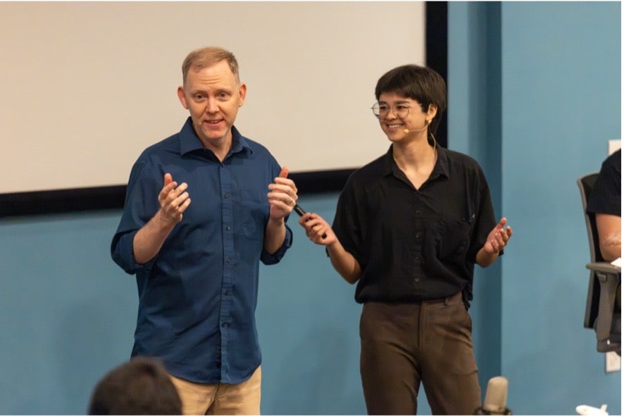 Two people stand in front of a screen giving a presentation; one gestures with hands while the other holds a remote and smiles.