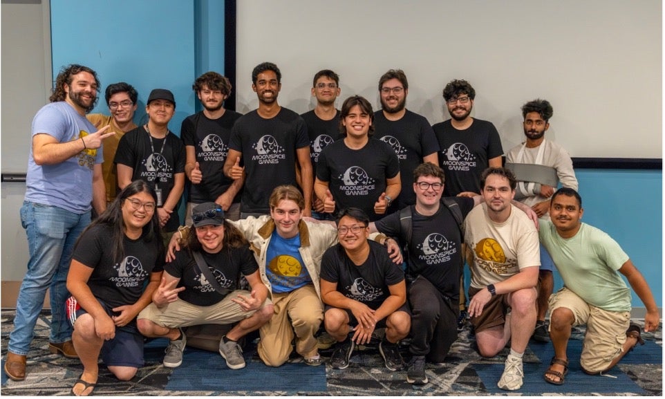 A group of seventeen people pose for a group photo indoors, with many wearing matching black "Moonshot Games" shirts and smiling at the camera.