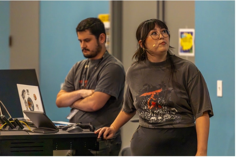 Two people stand at a podium. One looks at a screen with a headset on, while the other stands with arms crossed in front of a laptop with stickers.