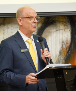 A man in a blue suit and yellow tie speaks into a microphone while holding papers at a podium, with abstract art in the background.