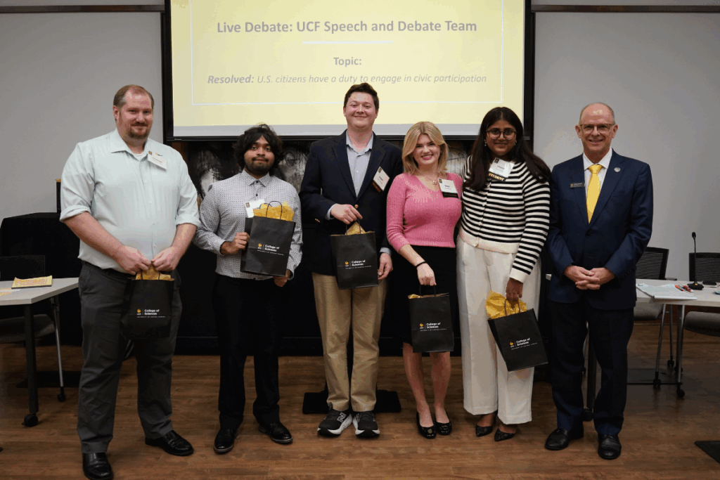 Six people stand in a row indoors, five holding gift bags. A screen behind them displays "Live Debate: UCF Speech and Debate Team" and the debate topic.