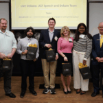 Six people stand in a row indoors, five holding gift bags. A screen behind them displays "Live Debate: UCF Speech and Debate Team" and the debate topic.