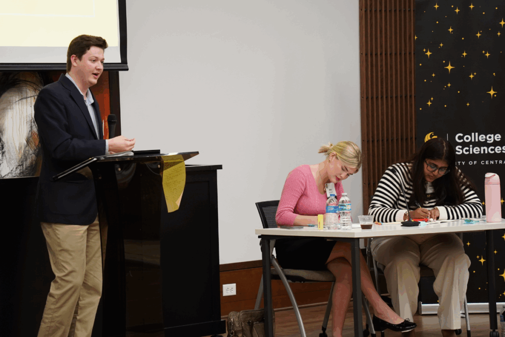 A young man speaks at a podium while two women sit at a table, one listening and the other writing notes, during an academic event.
