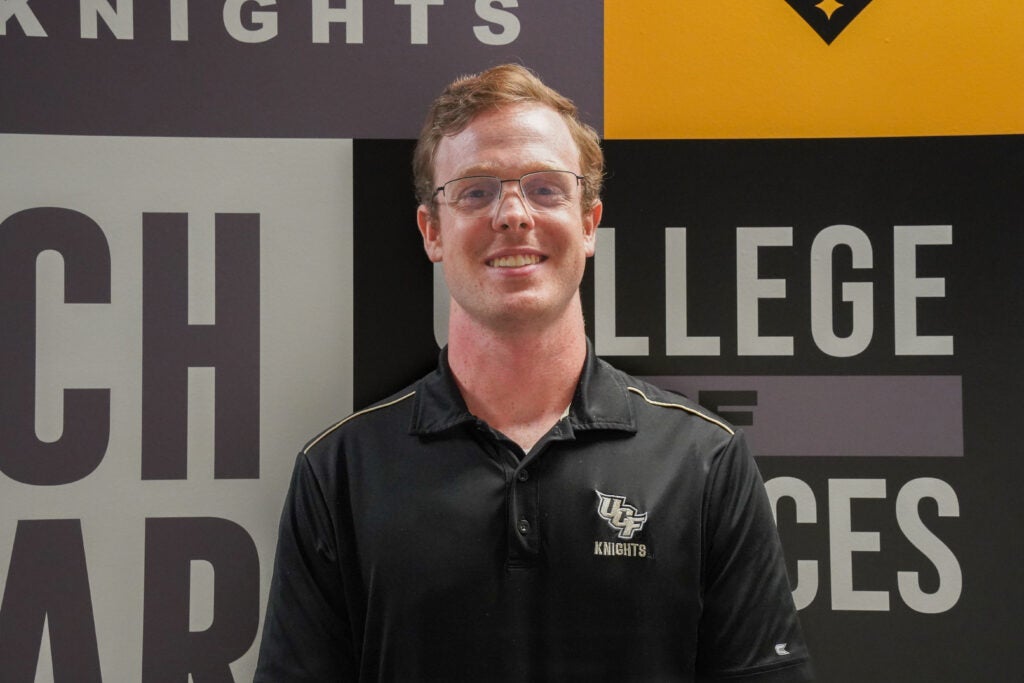 A person wearing glasses and a black UCF Knights polo stands in front of a wall with "COLLEGE OF SCIENCES" text and UCF branding.