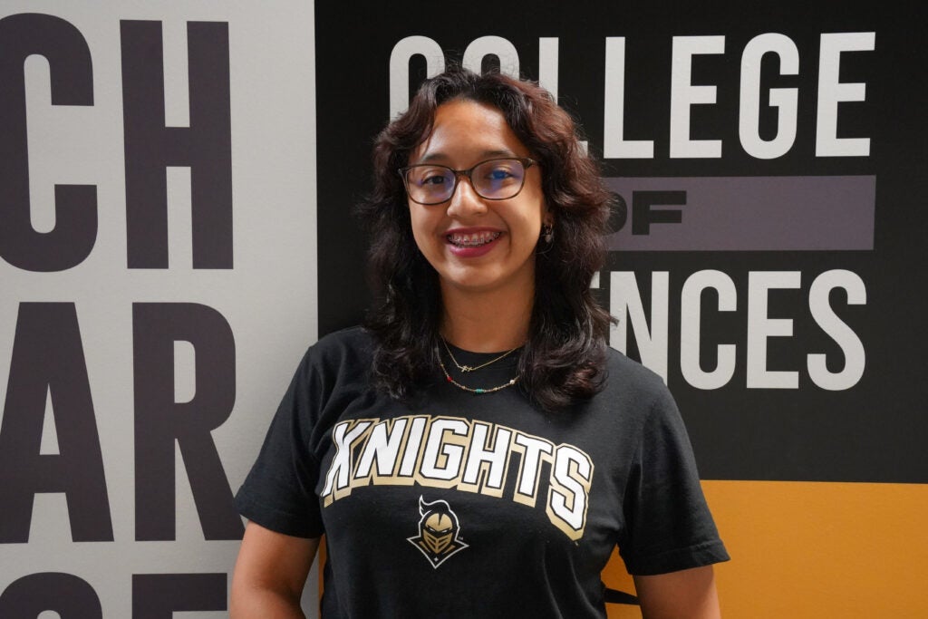 A person wearing glasses and a "Knights" t-shirt stands smiling in front of a wall with "College of Sciences" written on it.