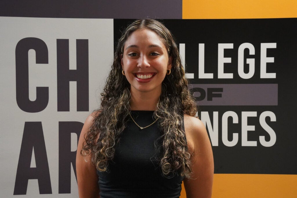 A woman with long curly hair and a black sleeveless top stands smiling in front of a "College of Arts and Sciences" sign.