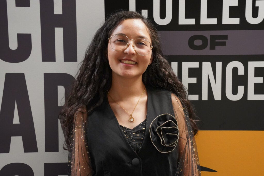 A young woman with long curly hair and glasses smiles while standing in front of a wall with the words "COLLEGE OF SCIENCE.