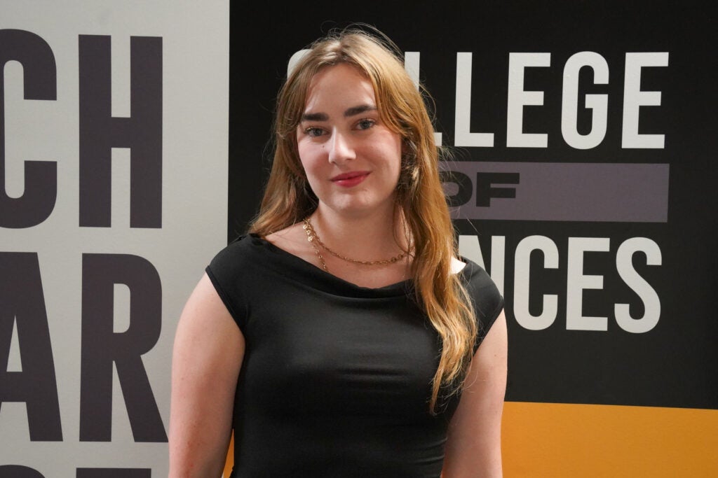 A woman with long light brown hair wearing a black dress stands in front of a wall with large text that reads "COLLEGE OF SCIENCES.