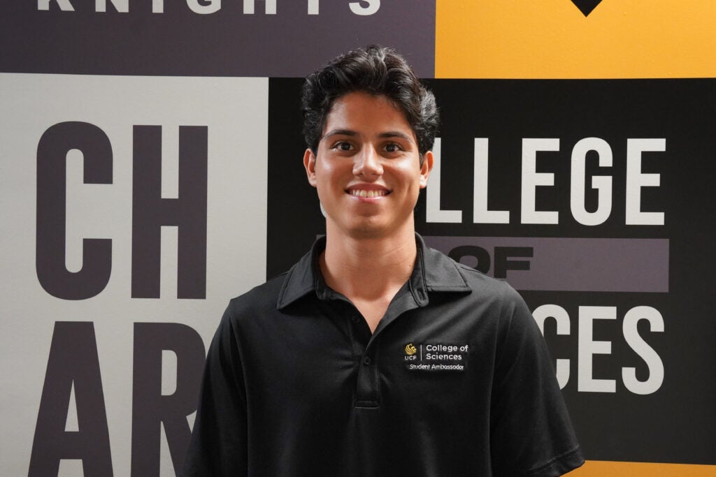 A young man wearing a black "College of Sciences" polo shirt stands smiling in front of a wall with bold, partially visible text.