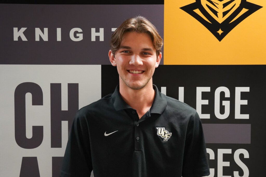 A young man in a black UCF polo shirt stands smiling in front of a wall with the words "Knights" and "College" and UCF logos.